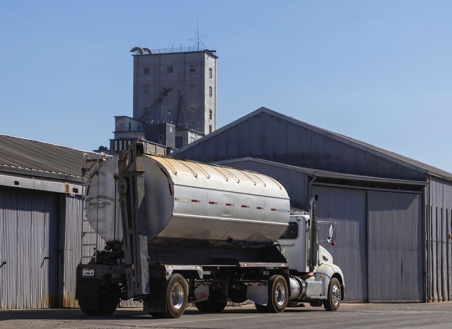 A large semi-trailer truck parked at an industrial grain warehouse under clear skies